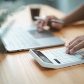 Hand eines kaufmännischen Angestellten, der einen Taschenrechner benutzt und etwas in ein Laptop eingibt.