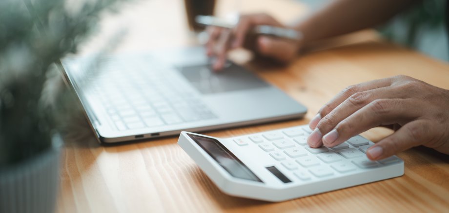 Hand eines kaufmännischen Angestellten, der einen Taschenrechner benutzt und etwas in ein Laptop eingibt.