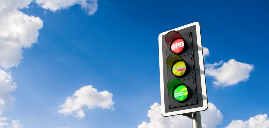 Ampel mit den Logos der drei Parteien SPD, FDP und Grüne vor einem leicht wolkigen Himmel.