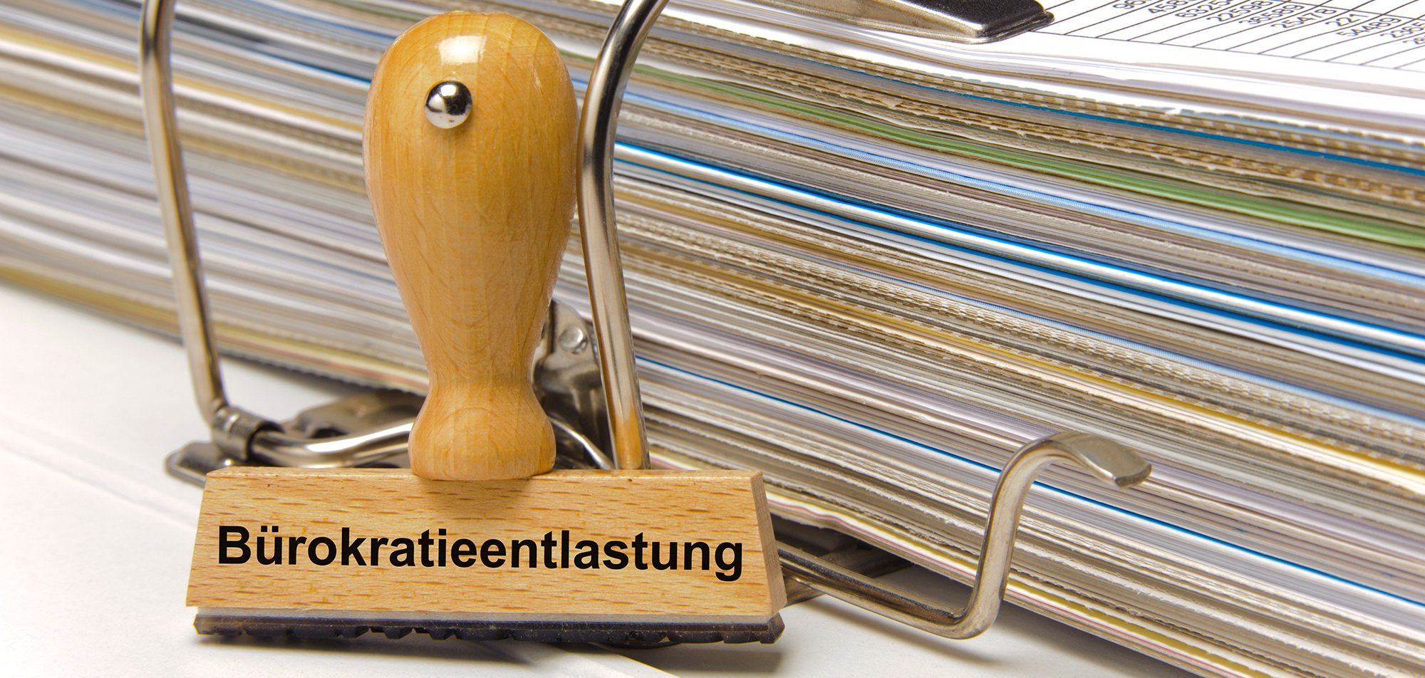 Ein Holzstempel ist beschriftet mit dem Aufdruck Bürokratieentlastung und lehnt an einem Aktenordner