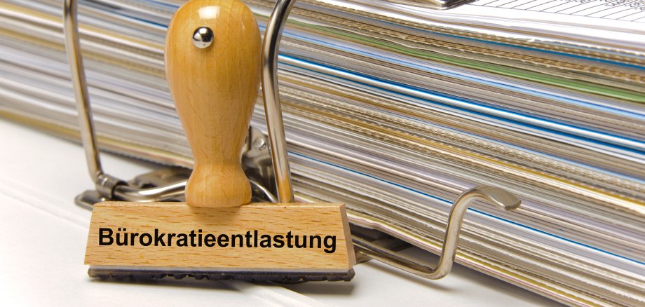 Ein Holzstempel ist beschriftet mit dem Aufdruck Bürokratieentlastung und lehnt an einem Aktenordner Ein Holzstempel ist beschriftet mit dem Aufdruck Bürokratieentlastung und lehnt an einem Aktenordner