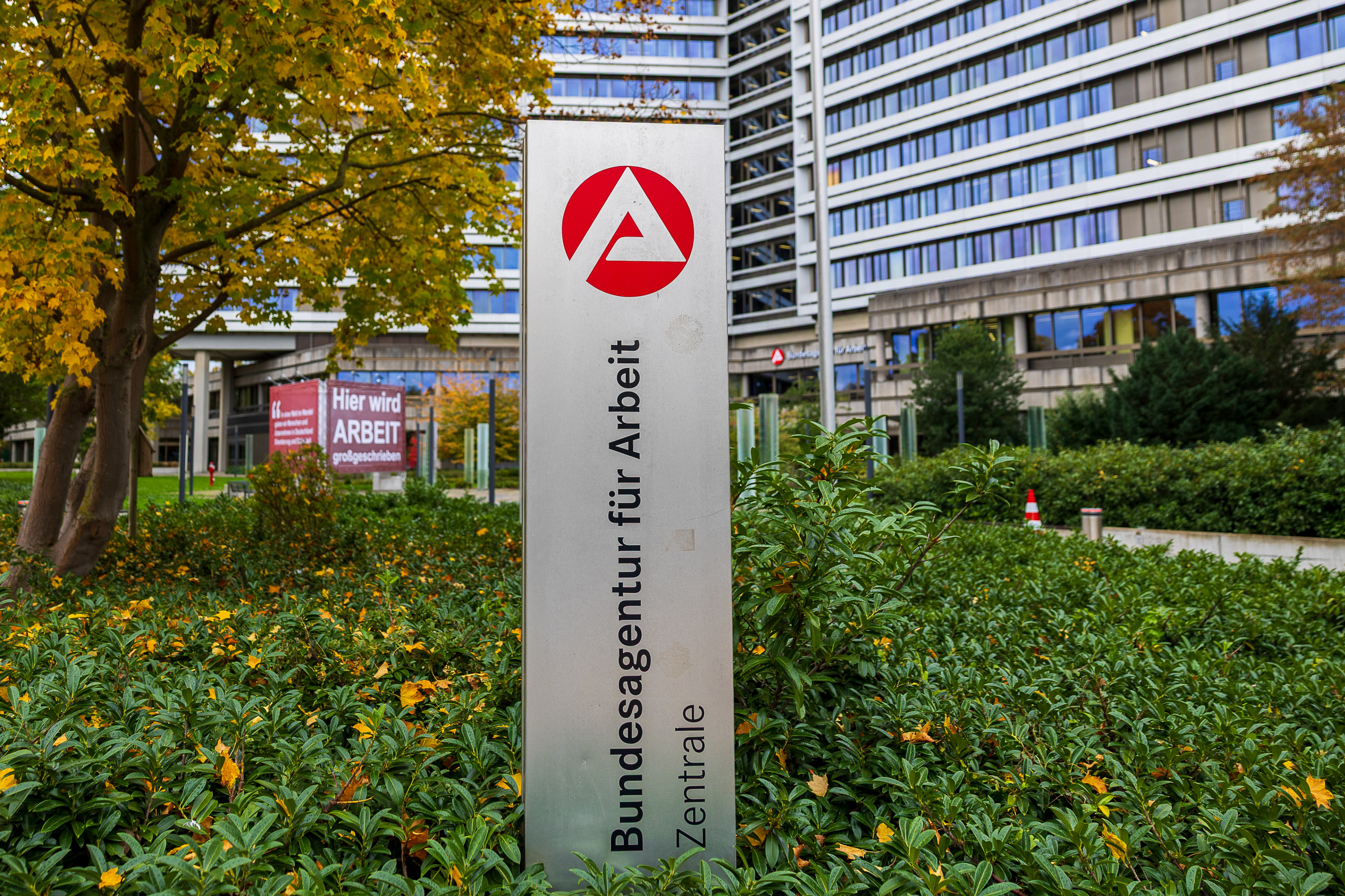Bundesagentur für Arbeit im Herbst, Nürnberg, 17.10.2025 Weitwinkelaufnahme der Zentrale der Bundesagentur für Arbeit in Nürnberg, Bayern, Deutschland. Im Vordergrund ist ein Hinweisschild mit dem Logo und Schriftzug der Behörde zu sehen. Im Hintergrund befinden sich das Hauptgebäude sowie zwei große Banner mit dem Slogan „Hier wird ARBEIT großgeschrieben“ und dem Leitbild der Institution. Die Szene ist von herbstlicher Bepflanzung eingerahmt.