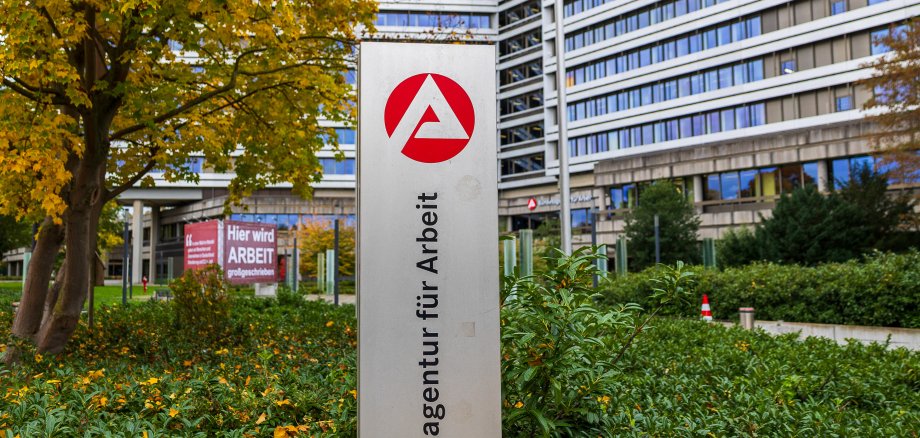 Weitwinkelaufnahme der Zentrale der Bundesagentur für Arbeit in Nürnberg, Bayern, Deutschland. Im Vordergrund ist ein Hinweisschild mit dem Logo und Schriftzug der Behörde zu sehen. Im Hintergrund befinden sich das Hauptgebäude sowie zwei große Banner mit dem Slogan „Hier wird ARBEIT großgeschrieben“ und dem Leitbild der Institution. Die Szene ist von herbstlicher Bepflanzung eingerahmt.