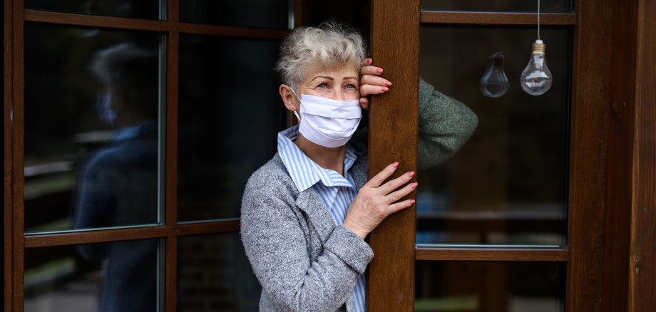 Traurige ältere Dame mit medizinischer Maske steht an ihrer Haustür Traurige ältere Dame mit medizinischer Maske steht an ihrer Haustür