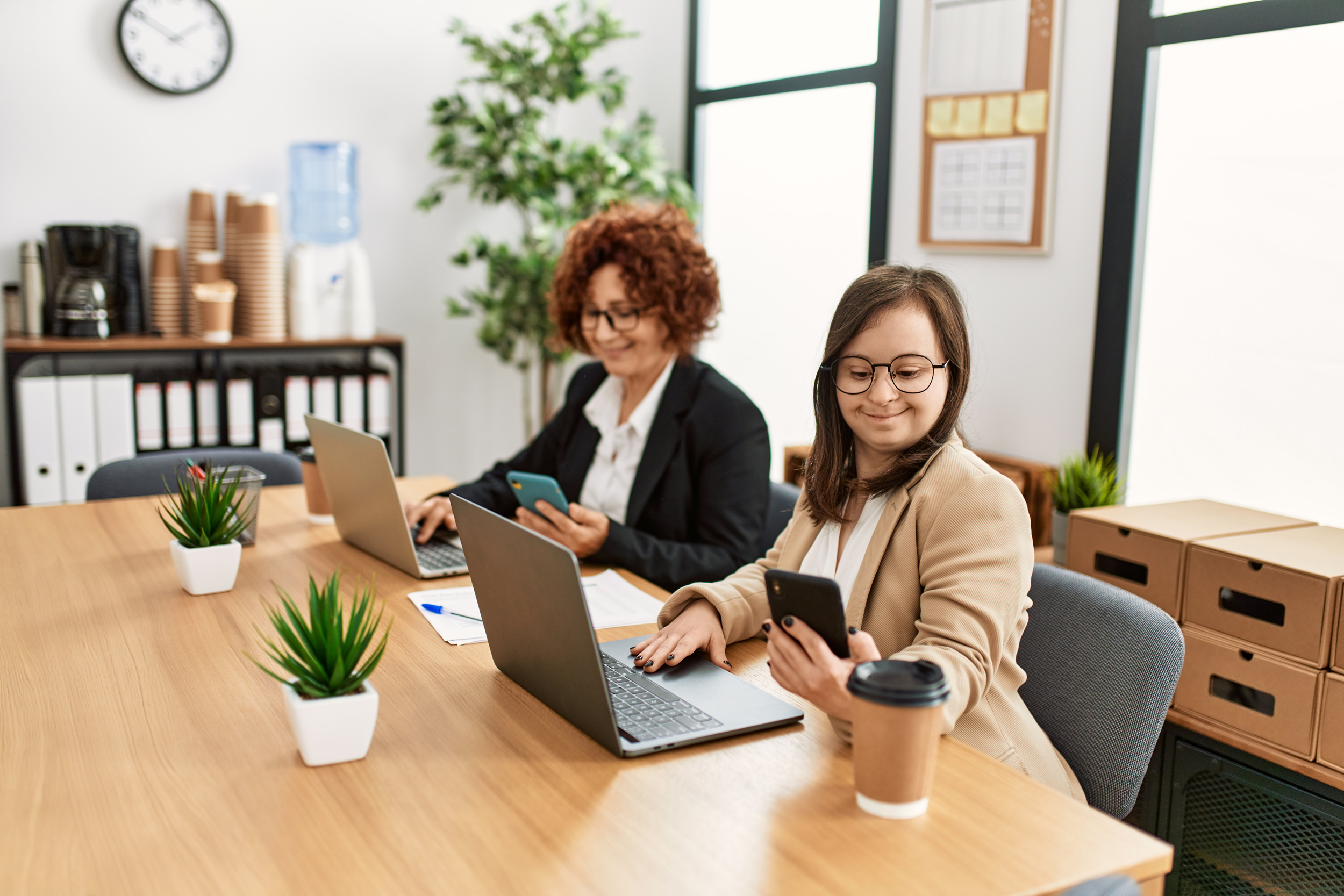 Zwei Frauen an einem Tisch bei der Arbeit mit Laptops. Inklusives Team mit einer jungen Frau mit Down Syndrom. 