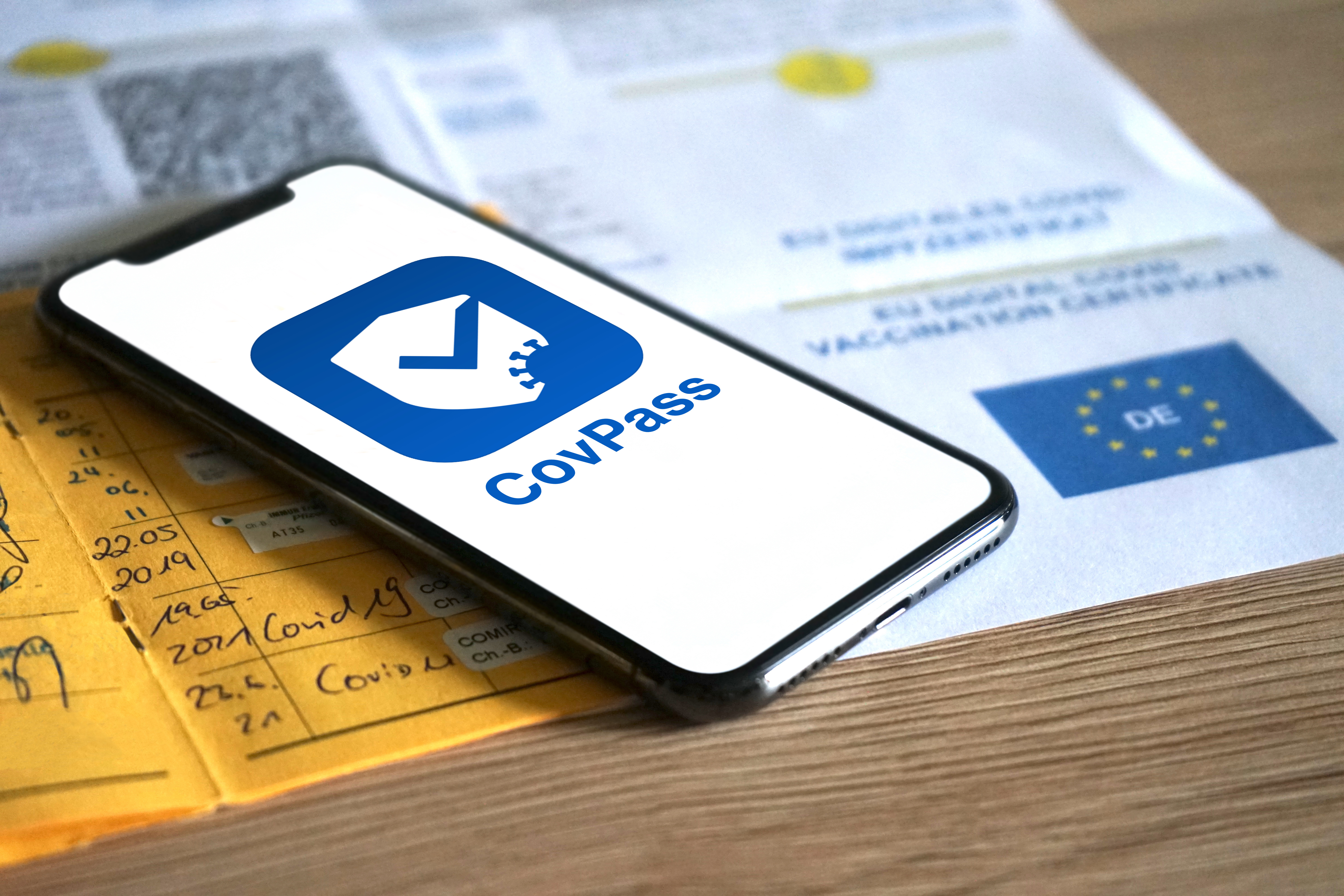 Smartphone mit Covpass App im Vordergrund. Dahinter unscharf  andere Impfnachweise wie  Ausdruck des Zertifikats und gelber Impfpass. 
