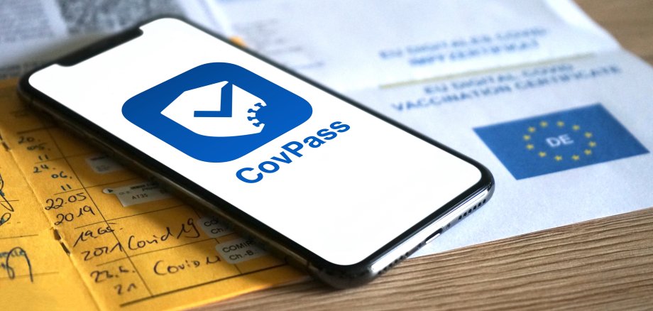 Smartphone mit Covpass App im Vordergrund. Dahinter unscharf andere Impfnachweise wie Ausdruck des Zertifikats und gelber Impfpass.
