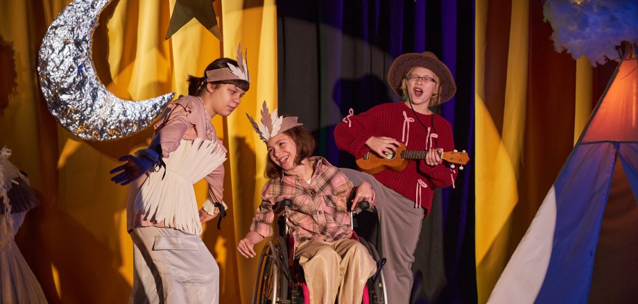 Ganzkörperaufnahme von drei fröhlichen jungen Sängerinnen, die auf der Bühne ein Country-Lied darbieten, an einem inklusiven Kinder-Theaterstück im Western-Stil teilnehmen und in einem hellen Scheinwerferlicht stehen.