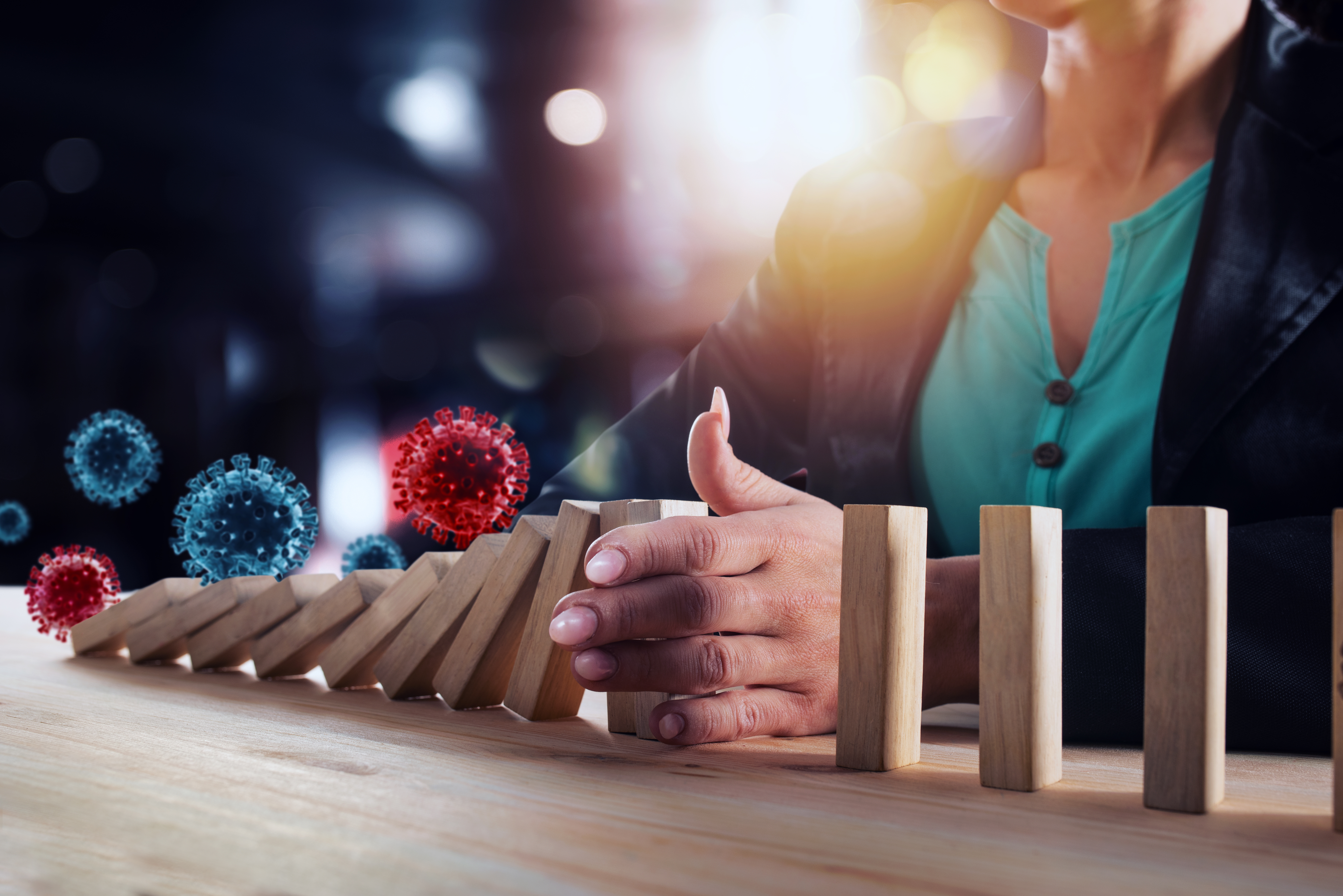 Businessfrau stoppt eine fallende Dominosteinreihe, über der Viren schweben. Ein Symbolbild für das Verhindern einer schweren Wirtschaftskrise durch Vireninfektionen. 