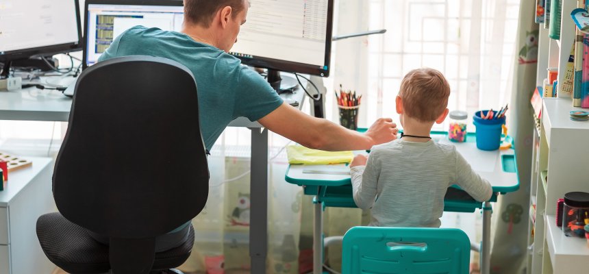 Vater, der während des  Corona-Lockdown zu Hause arbeitet, mit einem Kind, das nebendran an einem Kinderschreibtisch lernt. 