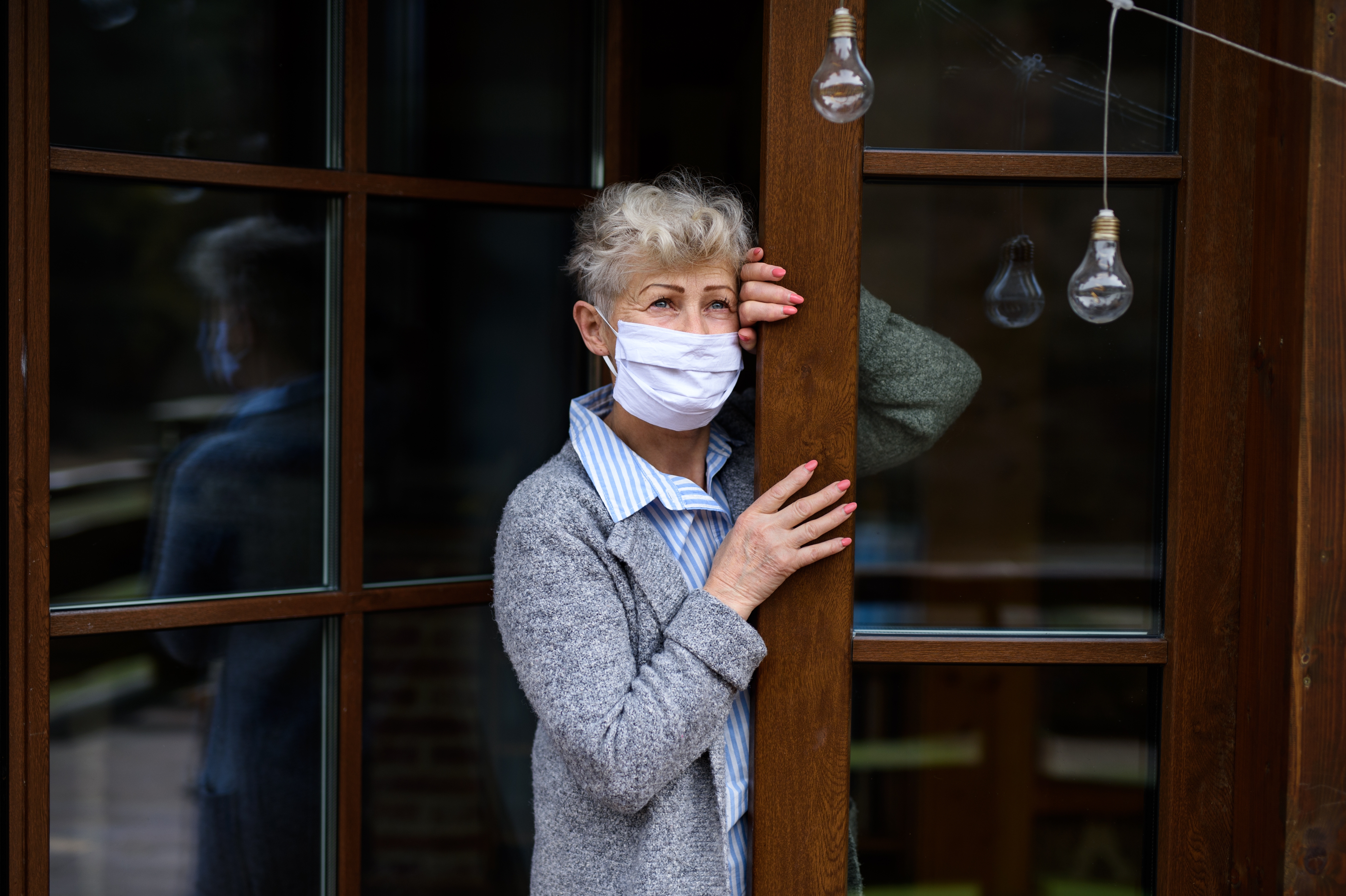 Traurige ältere Dame  mit medizinischer Maske steht an ihrer Haustür