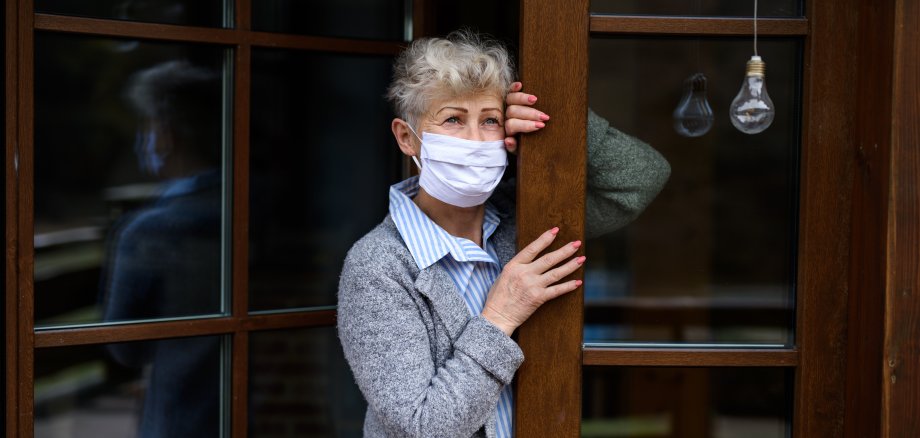 Traurige ältere Dame  mit medizinischer Maske steht an ihrer Haustür