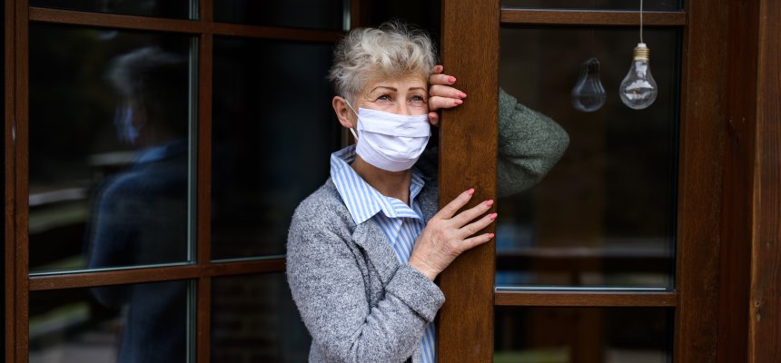 Traurige ältere Dame  mit medizinischer Maske steht an ihrer Haustür