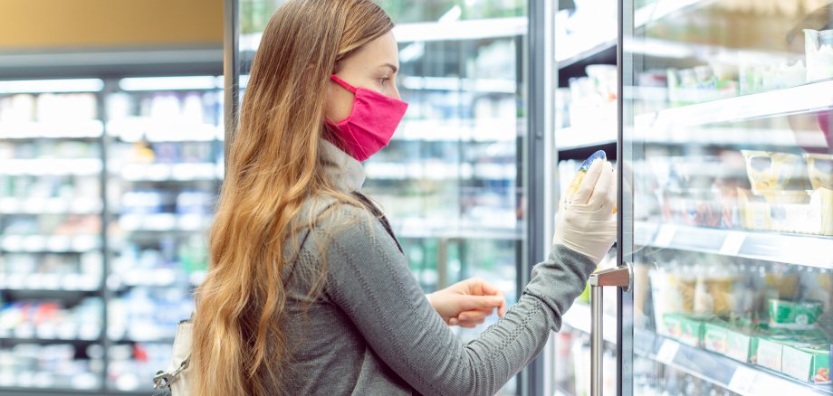 Frau mit medizinischer Maske im Supermarkt vor einem Frischeregal mit Türen.