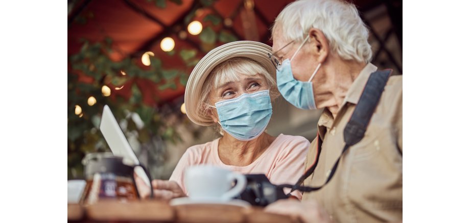 Zwei ältere Menschen sitzen in einem Straßencafé. Beide tragen eine medizinische Maske.