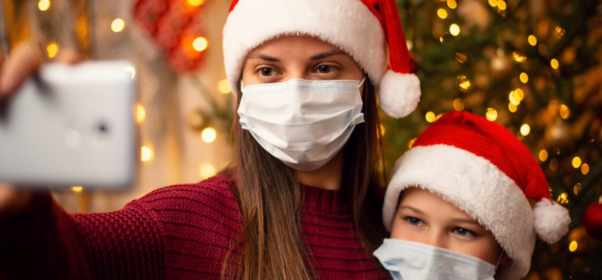 Mutter und Sohn machen ein Selfie vor dem Weihnachtsbaum. Beide haben Weihnachtsmützen auf und tragen eine medizinische Maske. 