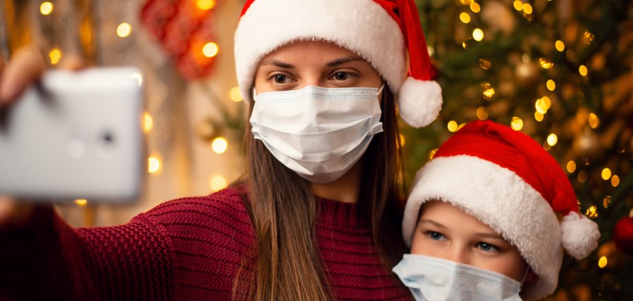 Mutter und Sohn machen ein Selfie vor dem Weihnachtsbaum. Beide haben Weihnachtsmützen auf und tragen eine medizinische Maske. 
