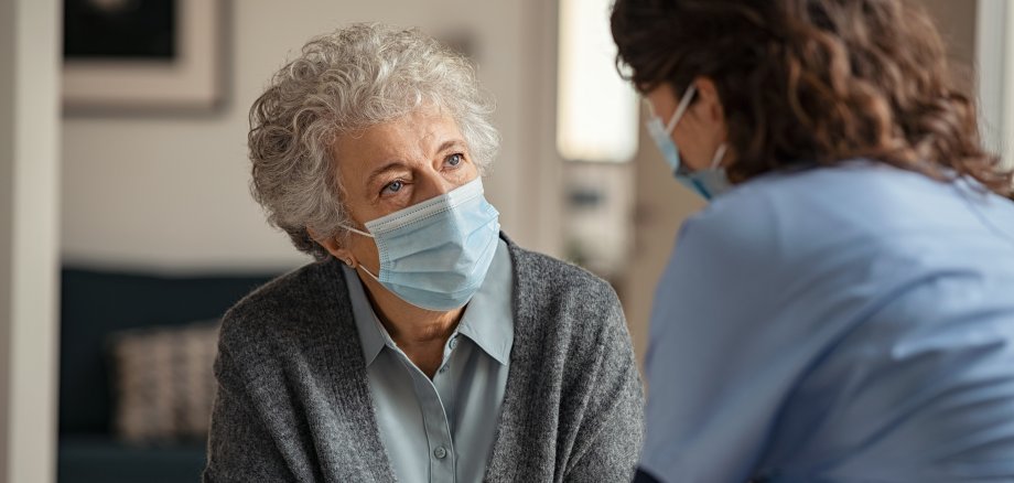 Ärztin, die mit einer älteren Patientin spricht. Beide tragen medizinische Masken.