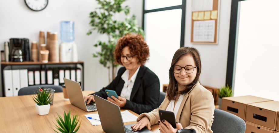 Zwei Frauen an einem Tisch bei der Arbeit mit Laptops. Inklusives Team mit einer jungen Frau mit Down Syndrom. 