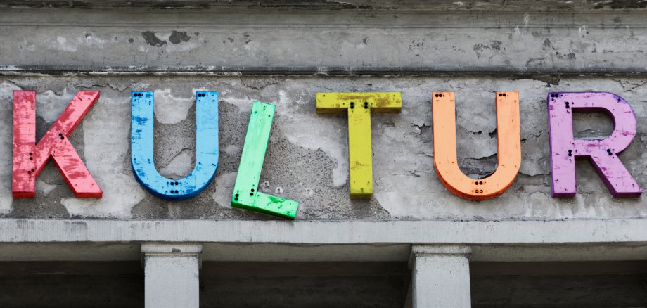Bunte Buchstaben, die ein wenig abgegriffen aussehen und draußen an einem Gebäude etwas schief angebracht sind, ergeben das Wort "Kultur".