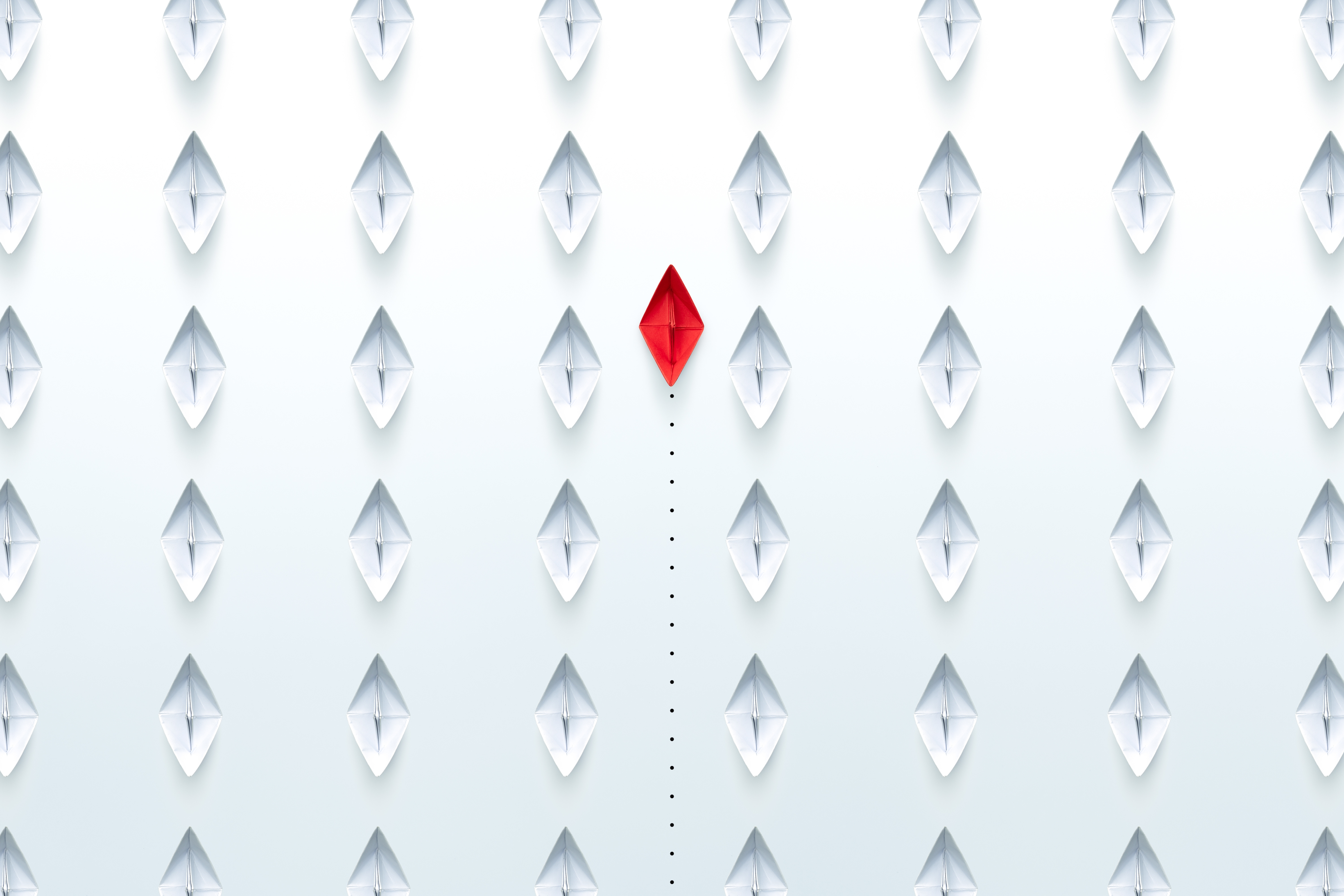 Weiße gefaltete Papierboote, gleichmässig auf einer weißen Fläche verteilt. In der Mitte befindet sich ein rotes gefaltetes Papierboot.